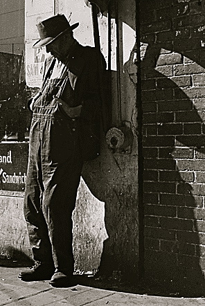 Man in overalls leaning against by old storefront. 1935 Dorothea Lange1(1895-1965)FDR Pres.Lib./ USPD: pub.date, by Fed employee (Commons.wikimedia.org)